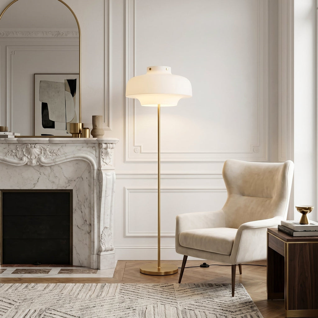 Beside a white marble fireplace and a cream wing chair, this Clérachic floor lamp glows softly, its brass stem rising from a round base.
