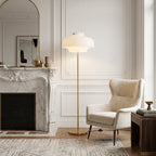 Beside a white marble fireplace and a cream wing chair, this Clérachic floor lamp glows softly, its brass stem rising from a round base.