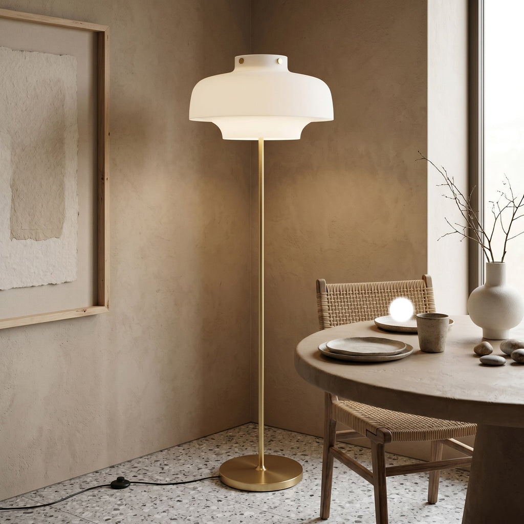 In a plaster-walled corner by a round wooden table, this Clérachic floor lamp stands on a speckled floor, its white shade lit warm.