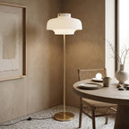 In a plaster-walled corner by a round wooden table, this Clérachic floor lamp stands on a speckled floor, its white shade lit warm.