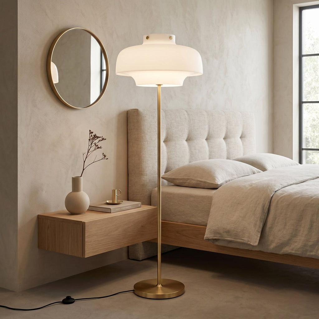 Beside a bed with a tufted headboard and a wooden bedside shelf, this Clérachic floor lamp shows its white shade and brass stem.