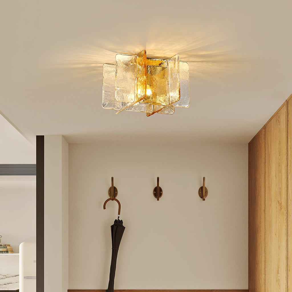 A Clérachic ceiling light with clear and amber glass panes glowing above an entrance wall with three brass wall hooks and an umbrella.