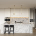 This Clérachic pendant light hangs over a white marble kitchen island, its frosted tube glowing softly beneath two thin gold rods.