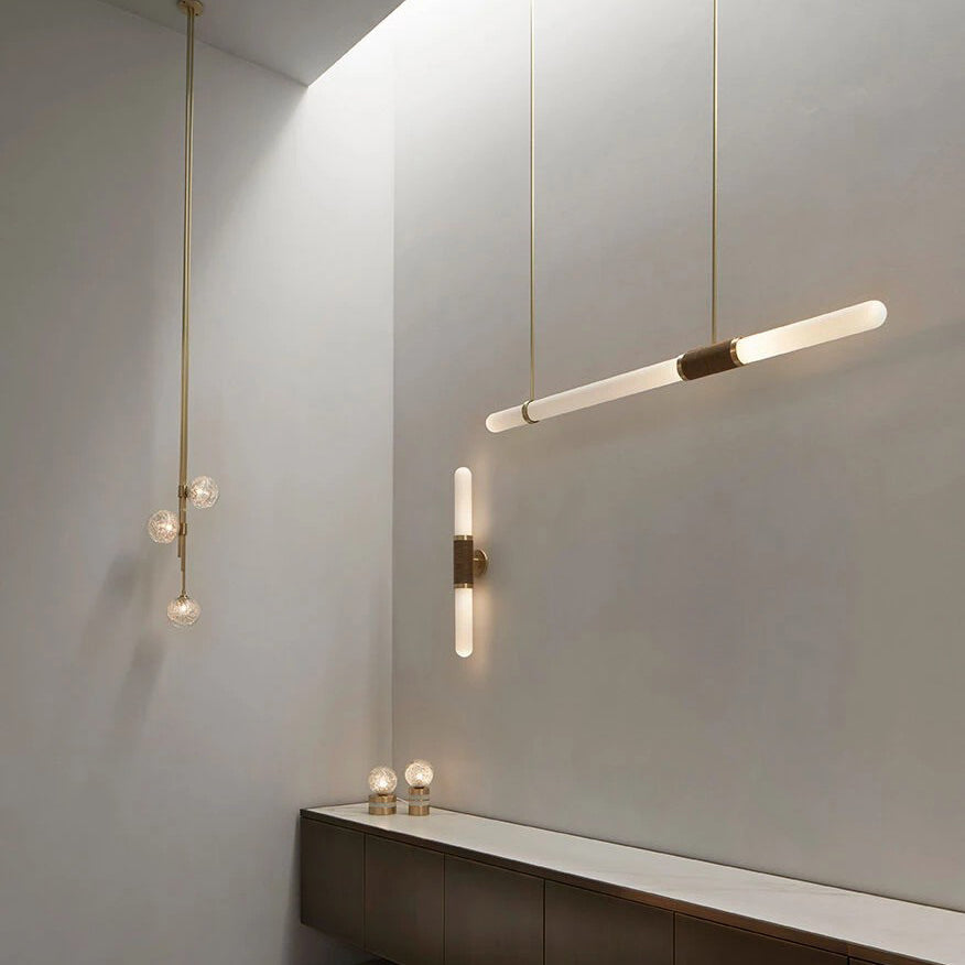 A Clérachic pendant light angled in a pale plaster room beside a lit wall light and a cluster of small glass globes on gold cords.