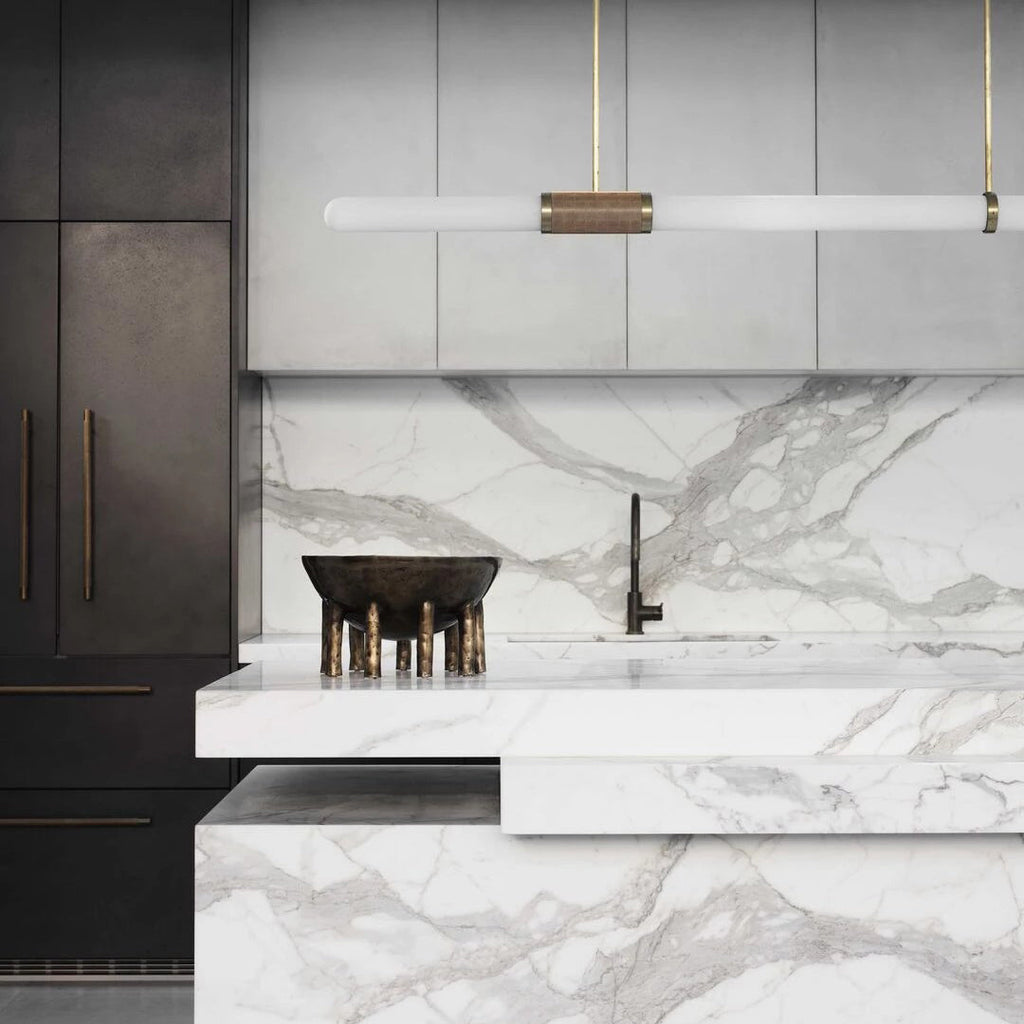 Close crop of this Clérachic pendant light above a white marble island, its gold-and-wicker centre band set against grey cabinetry.
