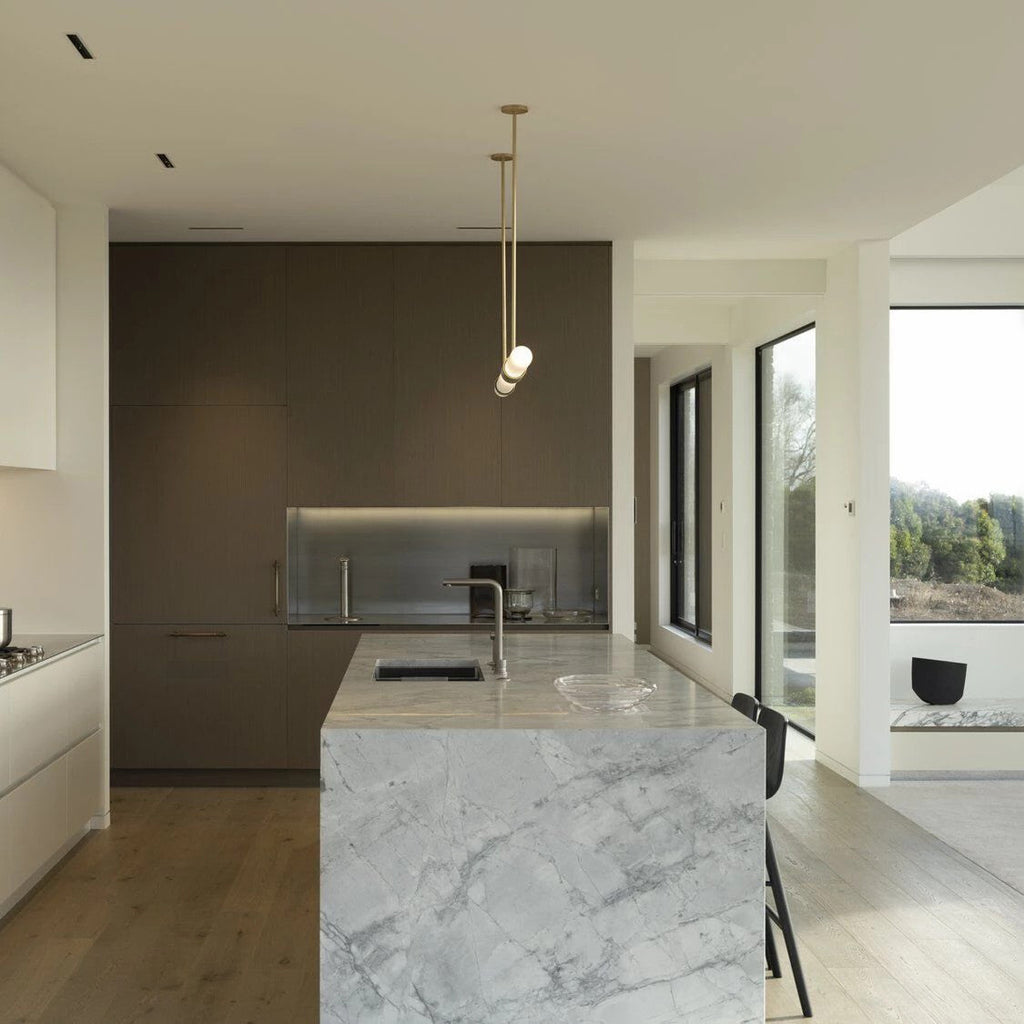A narrow vertical Clérachic pendant light hangs in a modern kitchen, with a stone-topped island and a dark cabinet wall behind.