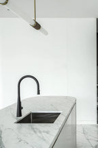 The end of this Clérachic pendant light enters the top of the frame above a marble island with a black pull-down tap.