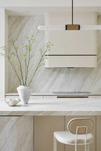 A Clérachic pendant light crosses the frame above a cream range hood, with a vase of fresh branches on the stone worktop below.