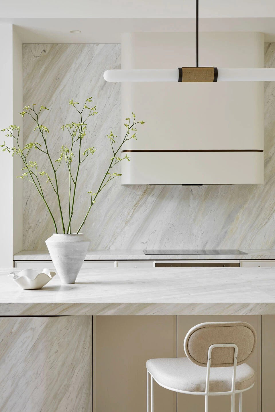 A Clérachic pendant light crosses the frame above a cream range hood, with a vase of fresh branches on the stone worktop below.
