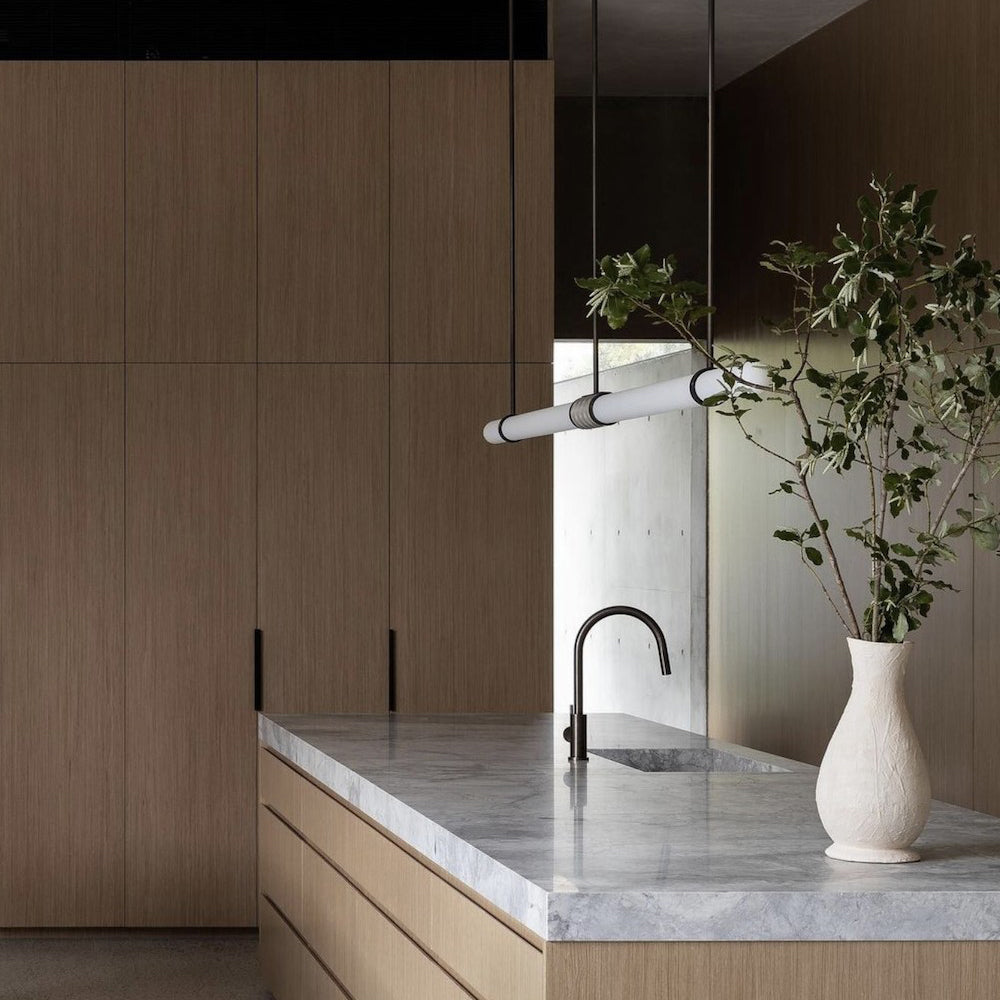 Angled view of this Clérachic pendant light beside a white vase of olive branches, with oak cabinets and a black tap behind.