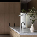 Angled view of this Clérachic pendant light beside a white vase of olive branches, with oak cabinets and a black tap behind.