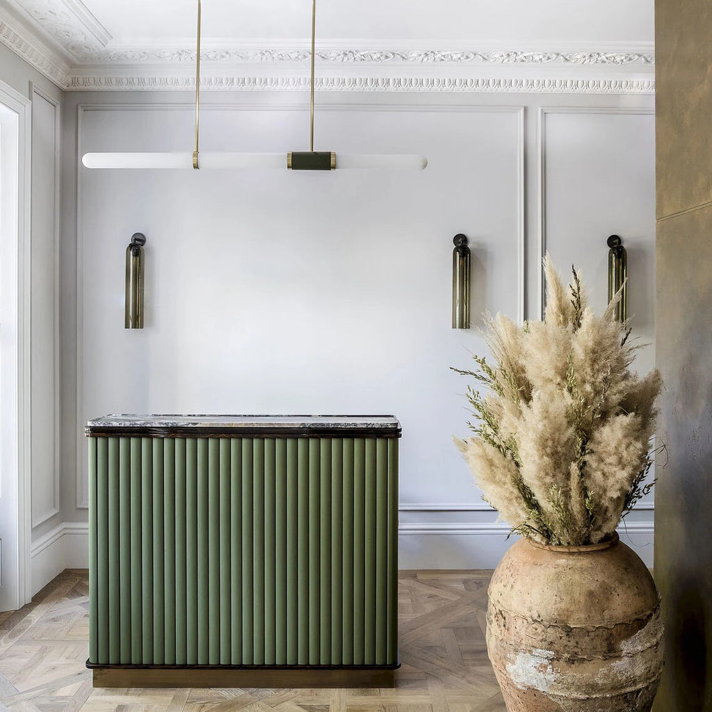 A Clérachic pendant light hangs in a panelled room with a green fluted island, wall lights and a tall jar of dried grasses.