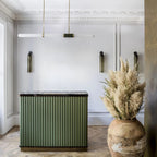 A Clérachic pendant light hangs in a panelled room with a green fluted island, wall lights and a tall jar of dried grasses.