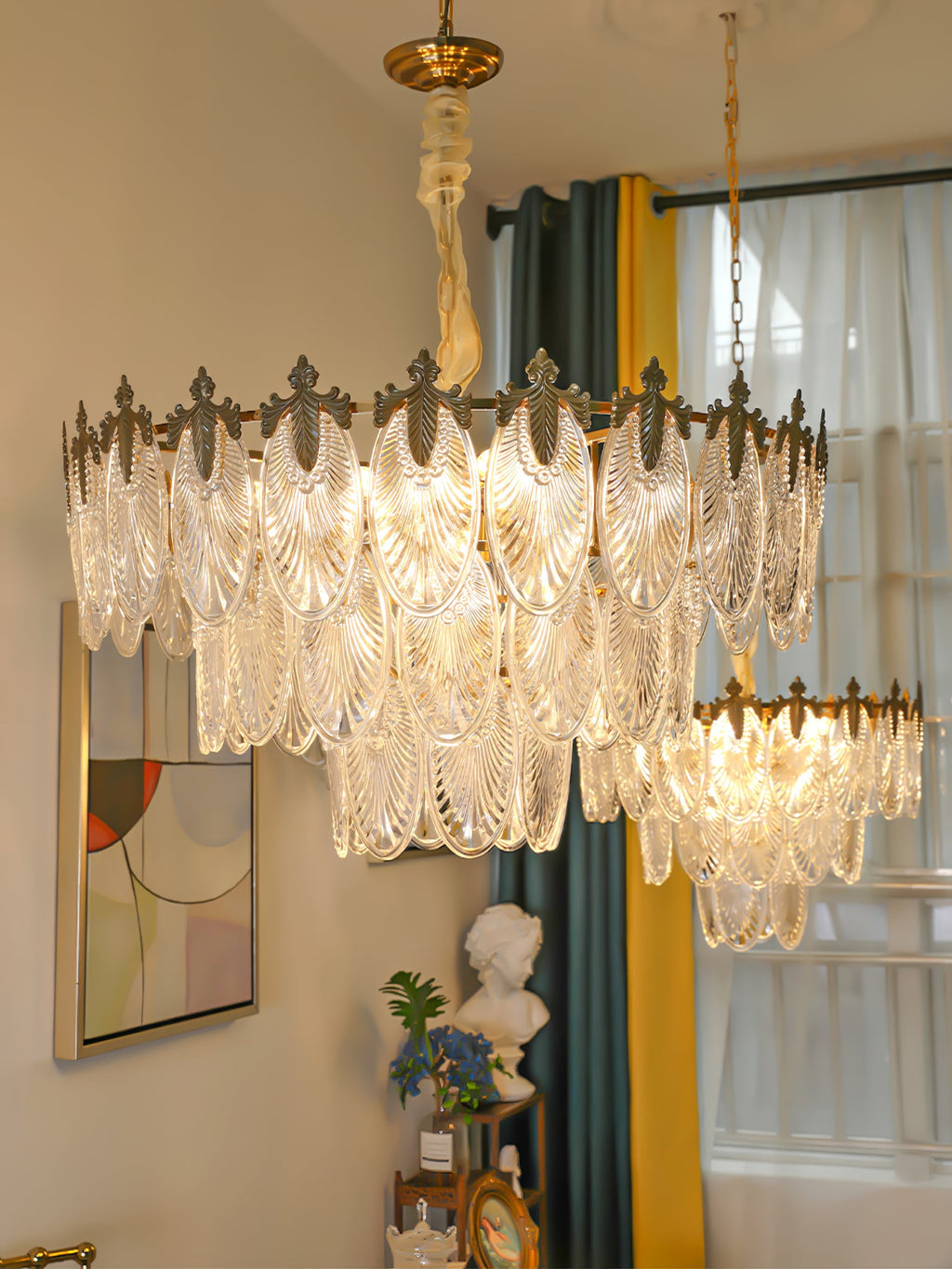 Two sizes of Clérachic glass leaf chandelier hang together in one room, with a teal and yellow curtain and a framed picture below.