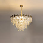 Warm light shining through the layered glass leaves of this Clérachic chandelier, with the gold crown at the top and a pale ceiling above.