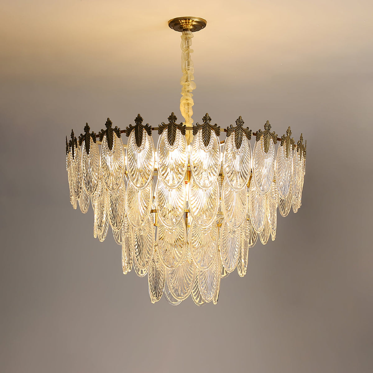 Large lit Clérachic chandelier with several tiers of glass leaves, hanging from a long chain against a warm grey backdrop.
