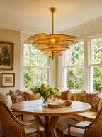 This Clérachic petal chandelier glows amber over a round wooden table in a breakfast nook, with tall windows and a wicker-backed bench.