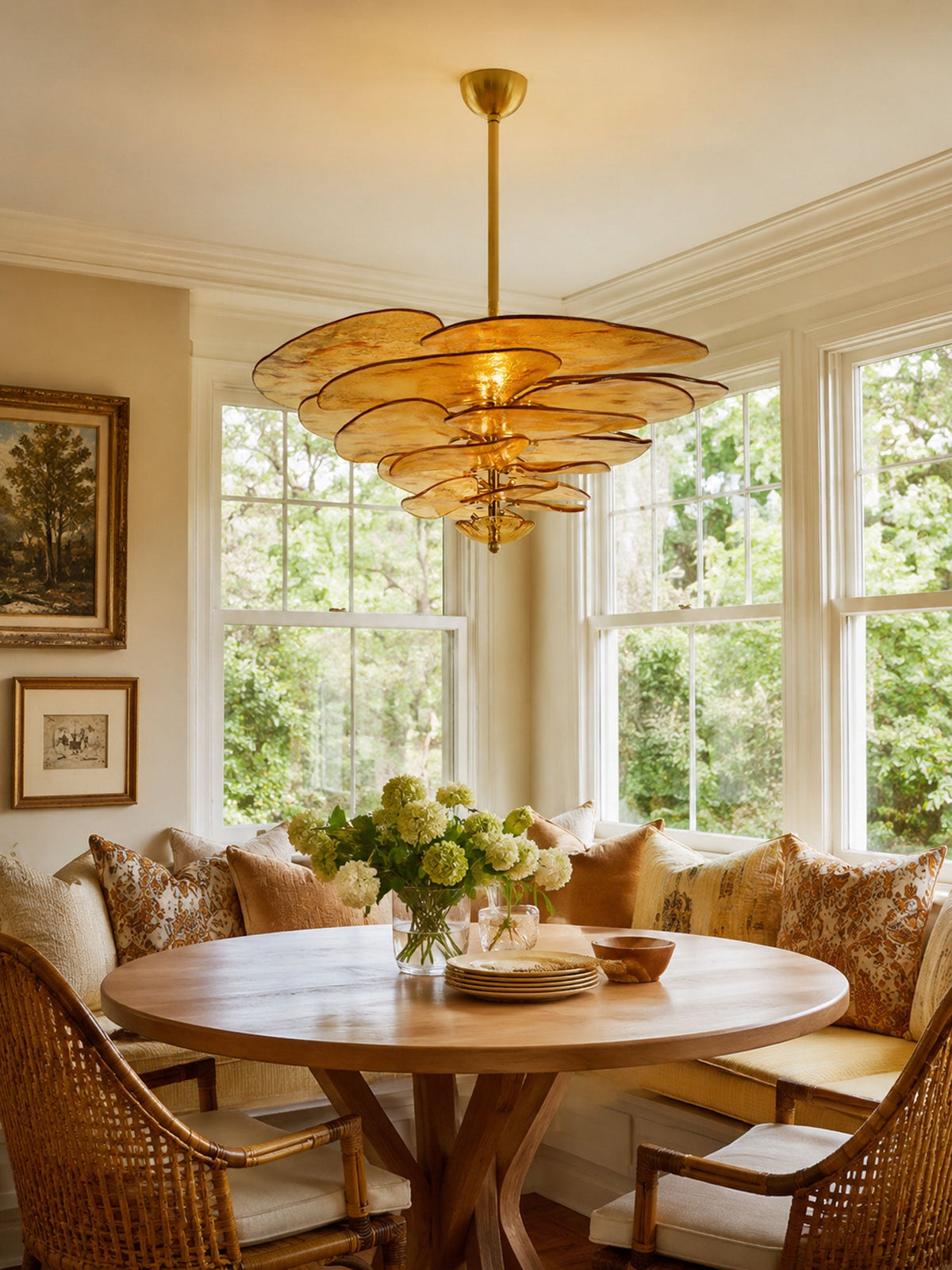 This Clérachic petal chandelier glows amber over a round wooden table in a breakfast nook, with tall windows and a wicker-backed bench.