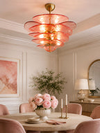 Pink Clérachic chandelier glowing in a pale room with a round table, pink velvet chairs and a bowl of peonies below.