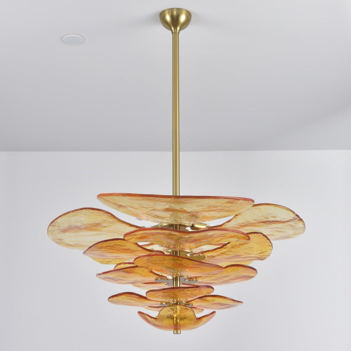 Amber Clérachic chandelier on a plain grey backdrop, its apricot-tinged glass petals stacked below a long brass rod.