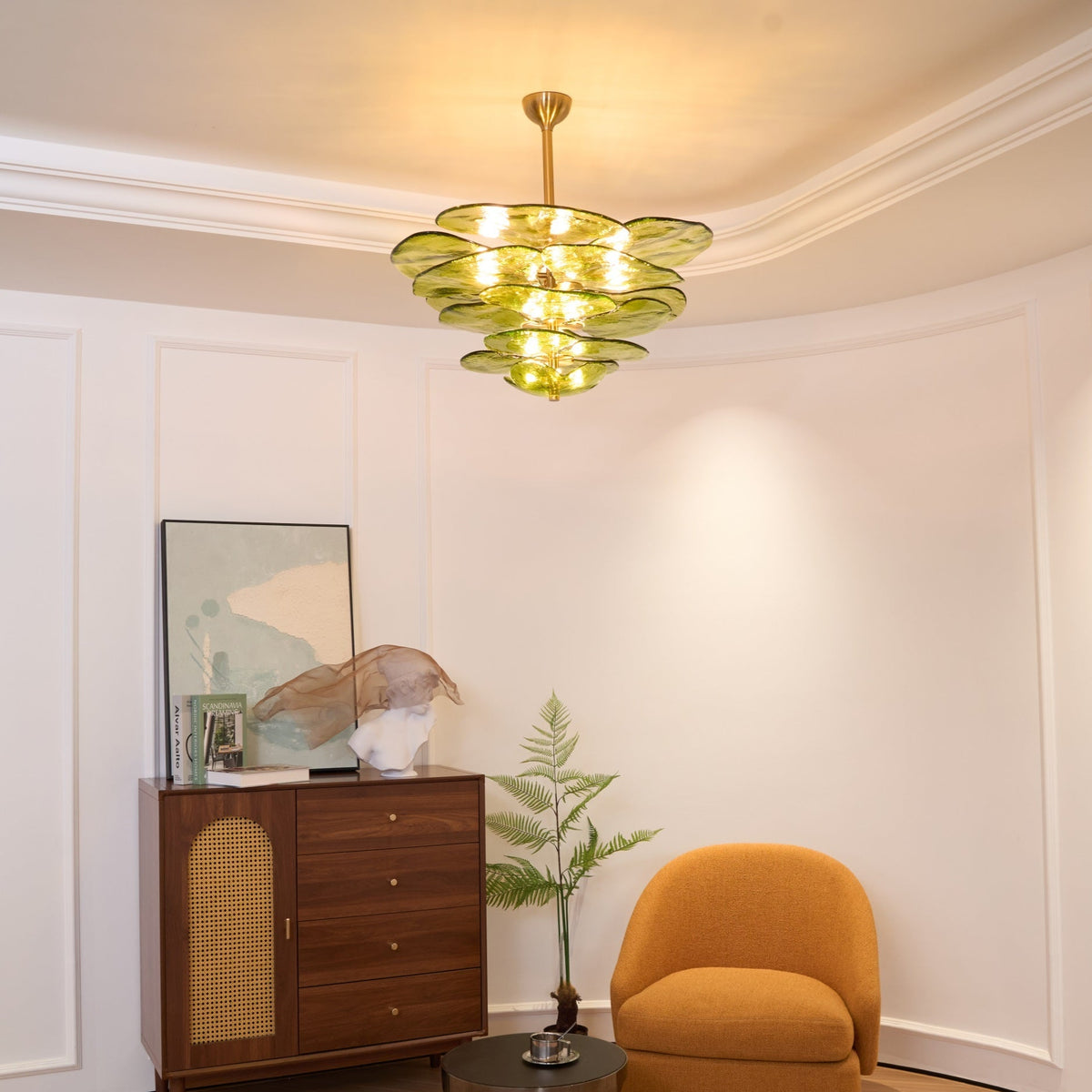 Small green Clérachic chandelier lit in a curved white room, above a wood cabinet with a cane front and a mustard armchair.