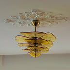 Amber Clérachic chandelier hung from a brass cup under an ornate white plaster ceiling rose, with a pale wall behind.