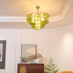 A lit green Clérachic chandelier hangs in a curved white room, above a wooden cabinet with a cane front and a leafy plant.