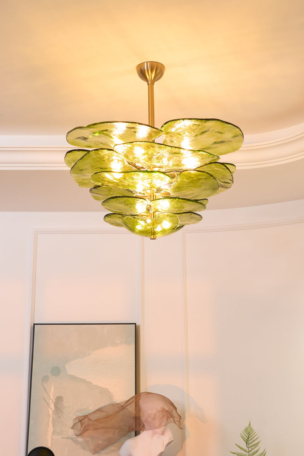 Narrow view of a lit green Clérachic chandelier below a coved ceiling, with a framed print and a sculpture beneath.