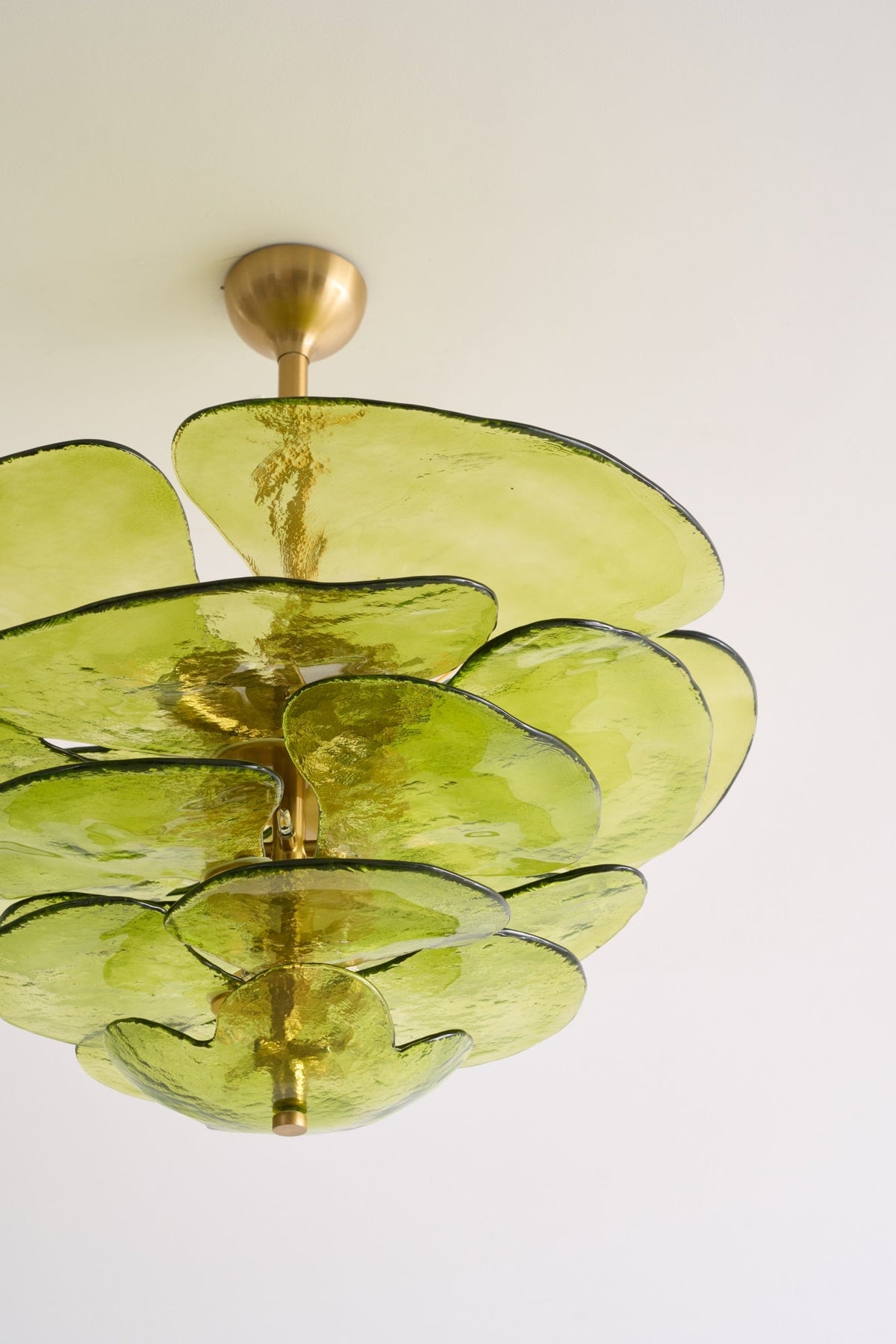 Close crop of the green glass petals and brass spindle on this Clérachic chandelier, lit by daylight against a white ceiling.