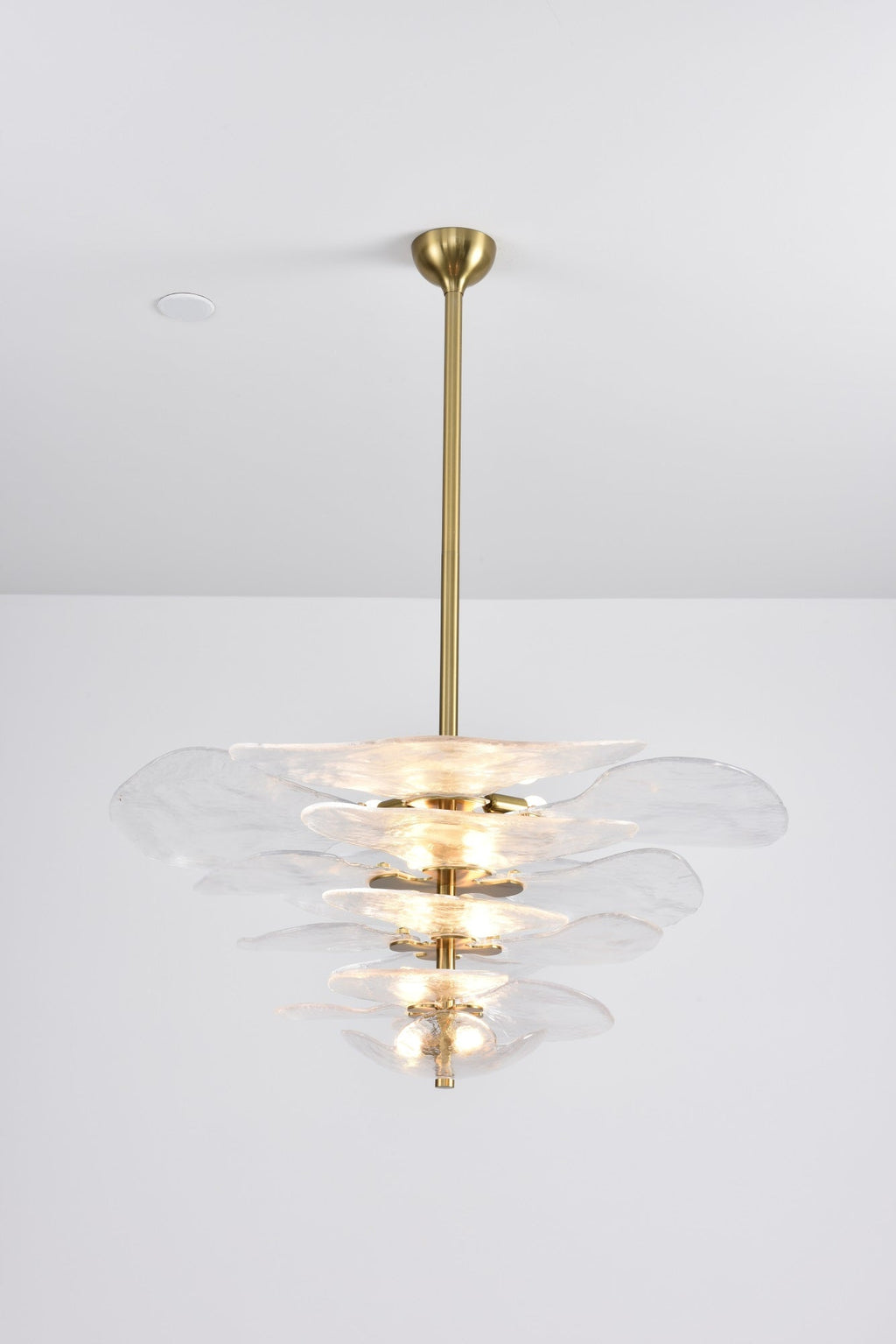 Lit clear glass Clérachic chandelier on a long brass rod, with a bright glow at its centre against a pale grey backdrop.