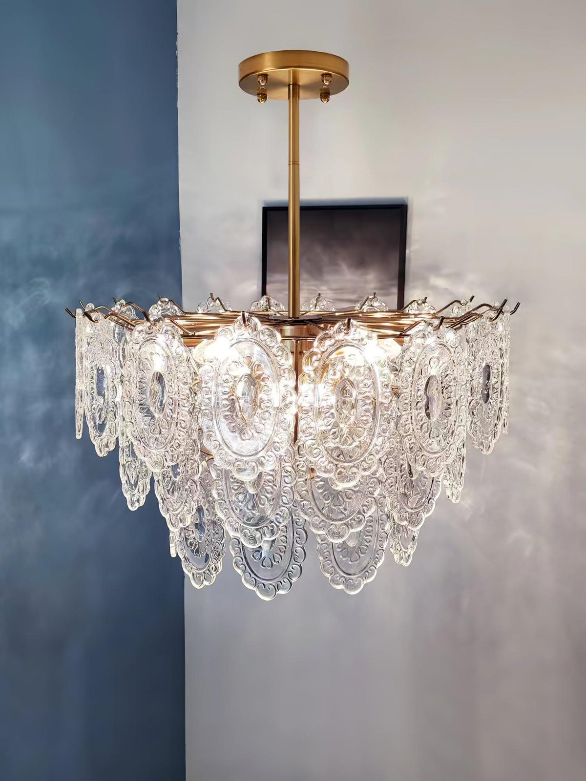 Clérachic glass pendant on a gold rod, lit against a blue-grey plastered wall, with a small dark frame behind it.