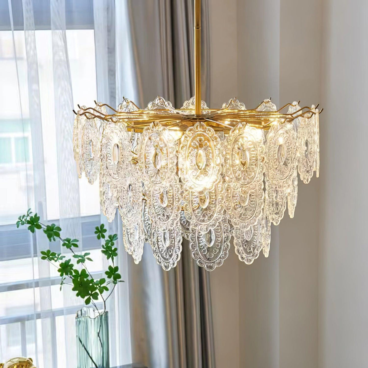 Clérachic glass pendant lit in front of a window with white sheers, a green plant and a gold wire ring at the top.