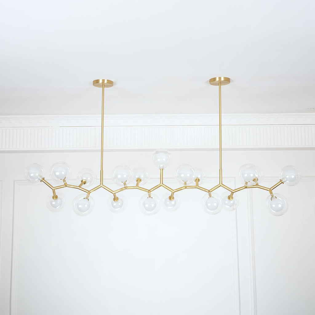 Unlit Clérachic chandelier with clear glass globes on a zigzag gold frame, hung from two rods against a white panelled wall.