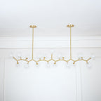 Unlit Clérachic chandelier with clear glass globes on a zigzag gold frame, hung from two rods against a white panelled wall.