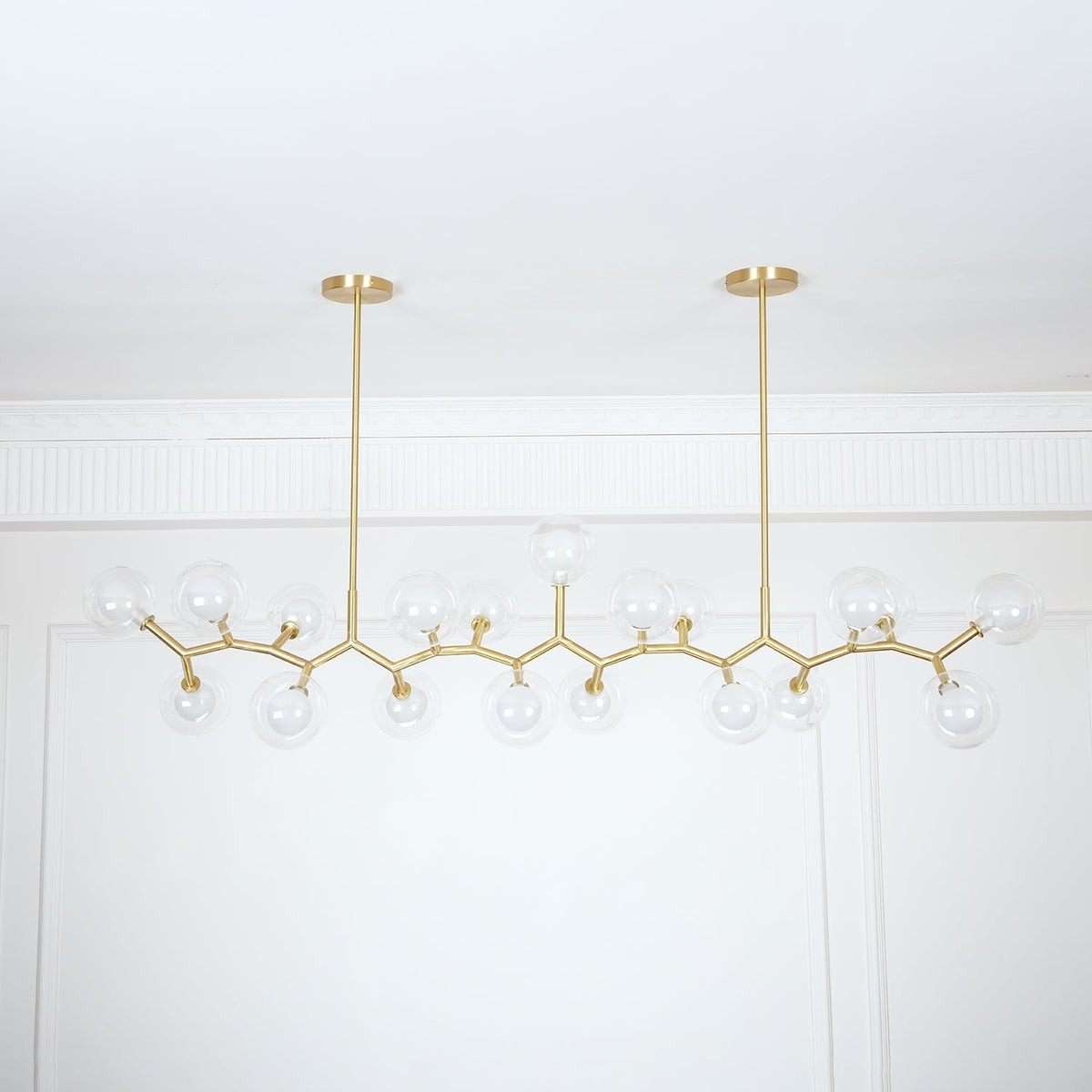 Unlit Clérachic chandelier with clear glass globes on a zigzag gold frame, hung from two rods against a white panelled wall.