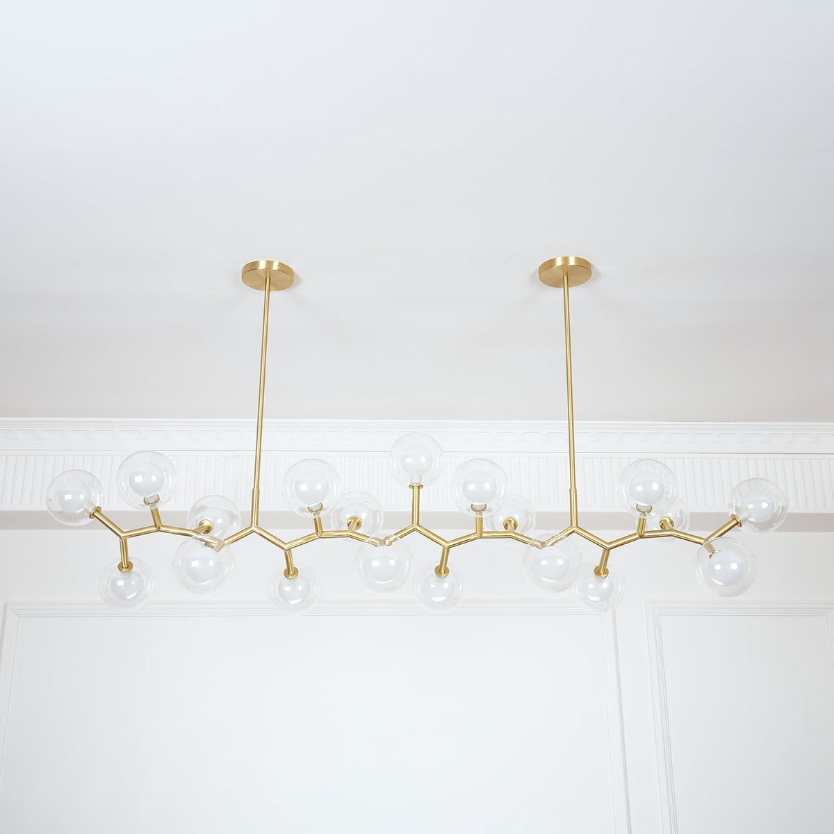 Unlit Clérachic chandelier with clear globes on thin gold branches, seen from slightly below under a white cornice.