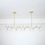 Wide view of the unlit Clérachic chandelier, its clear glass globes spread along gold branches under two slim hanging rods.
