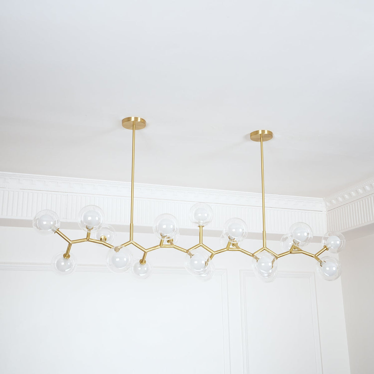 Clérachic chandelier with clear glass globes on gold branches, seen unlit beside a white panelled wall and ceiling moulding.