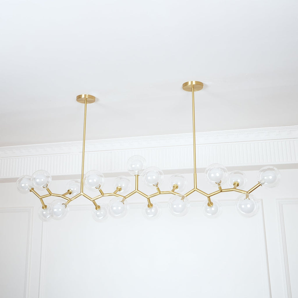 Unlit Clérachic chandelier on two gold rods, its clear glass globes spread out in a long line before a white panelled wall.