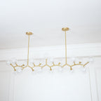 Unlit Clérachic chandelier on two gold rods, its clear glass globes spread out in a long line before a white panelled wall.
