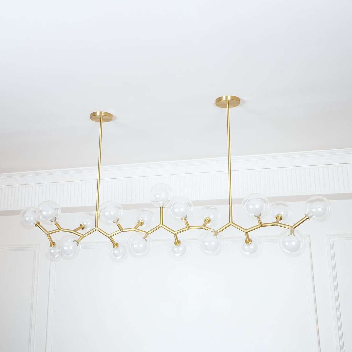 Unlit Clérachic chandelier on two gold rods, its clear glass globes spread out in a long line before a white panelled wall.
