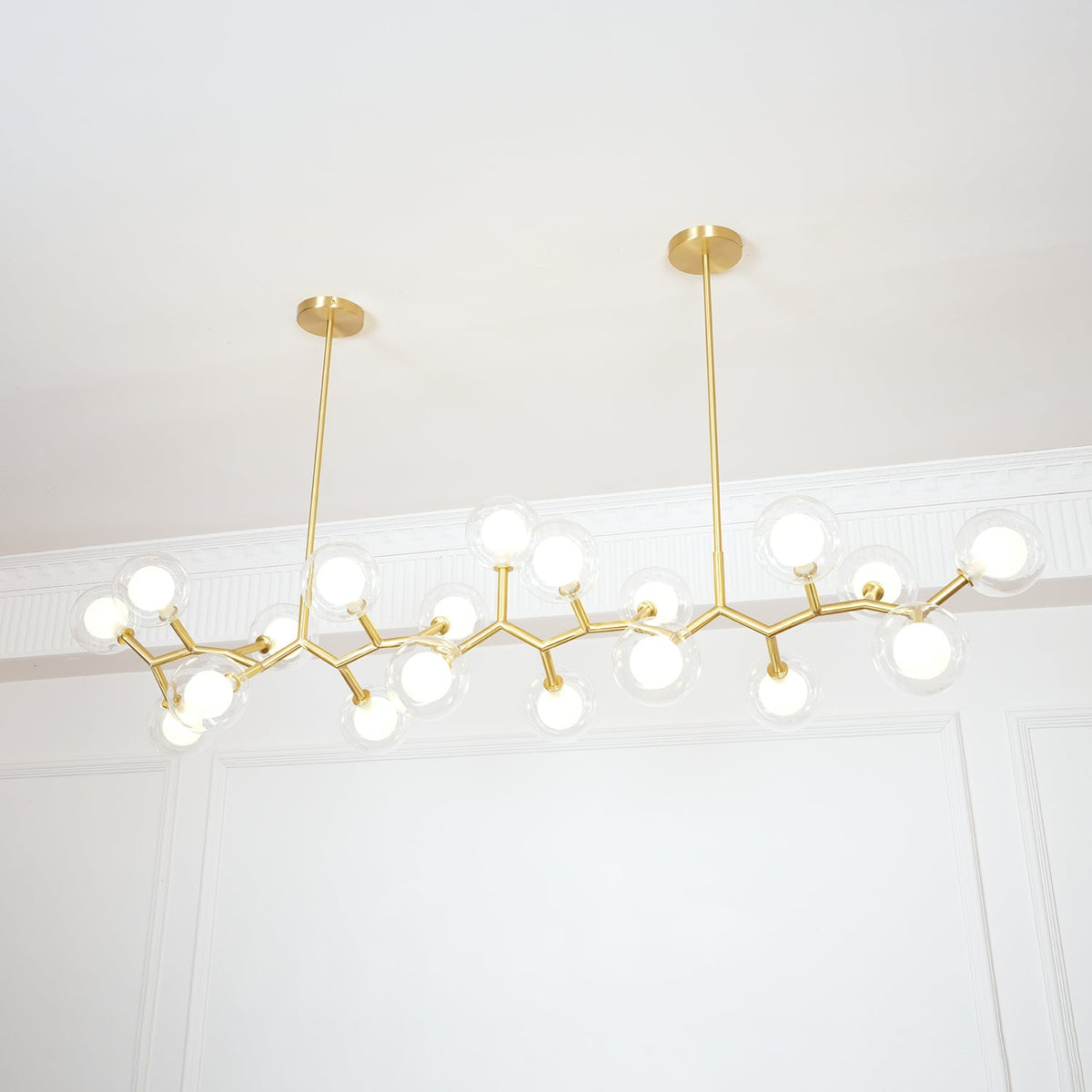 Lit Clérachic chandelier from below, its glowing globes and gold branches hung from two rods against white wall panelling.