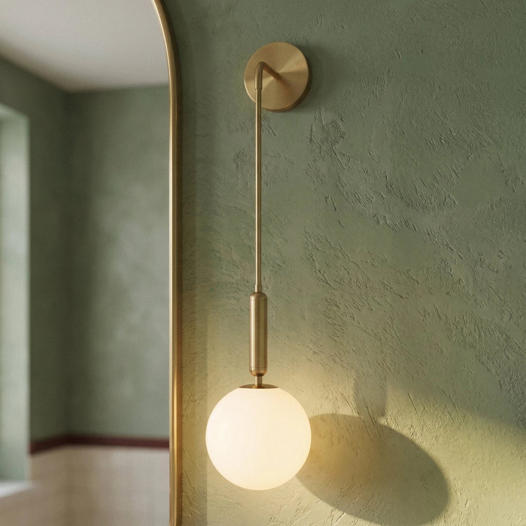 Close view of a Clérachic gold globe sconce's brass arm and opal glass globe against a sage green plaster wall.