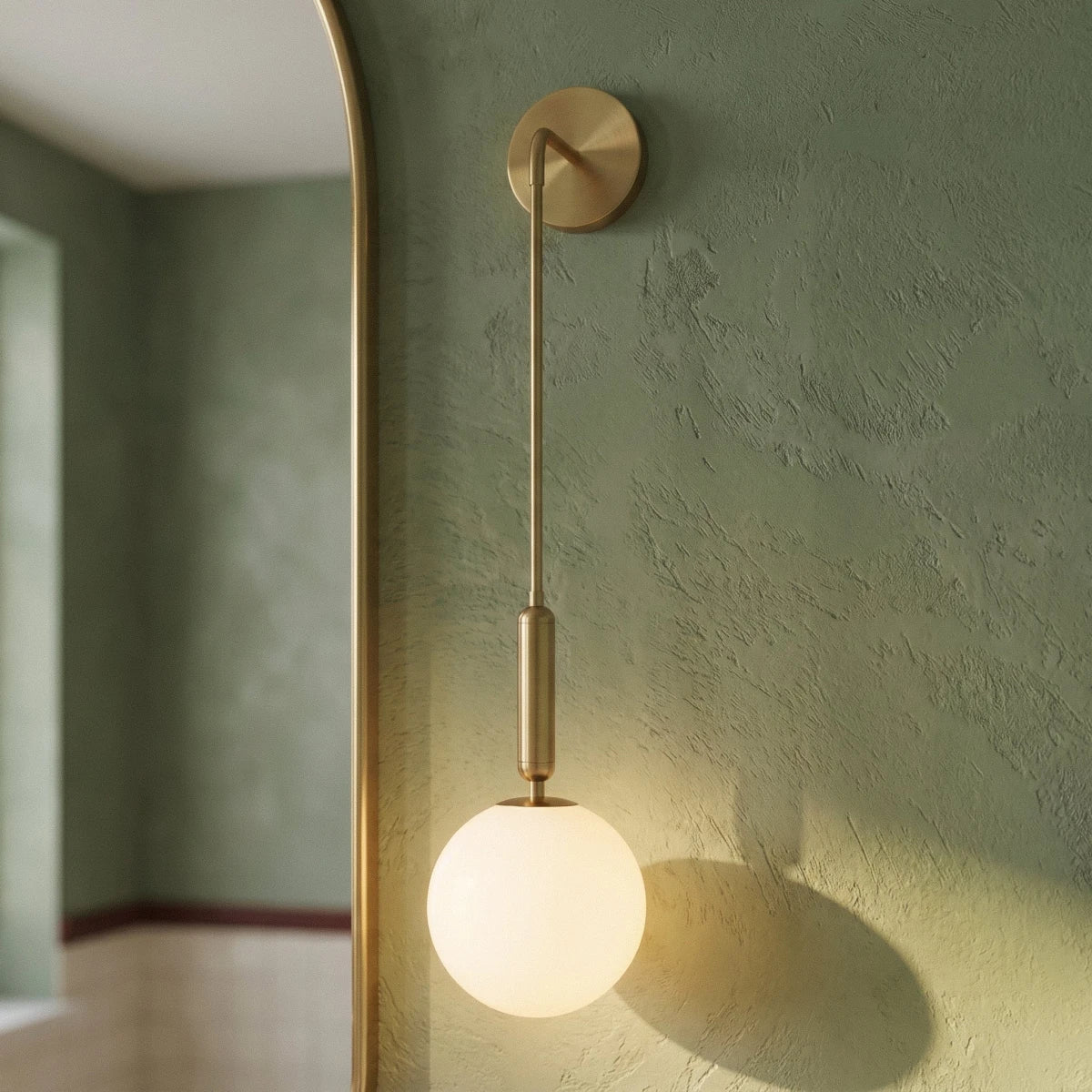 Close view of a Clérachic gold globe sconce's brass arm and opal glass globe against a sage green plaster wall.