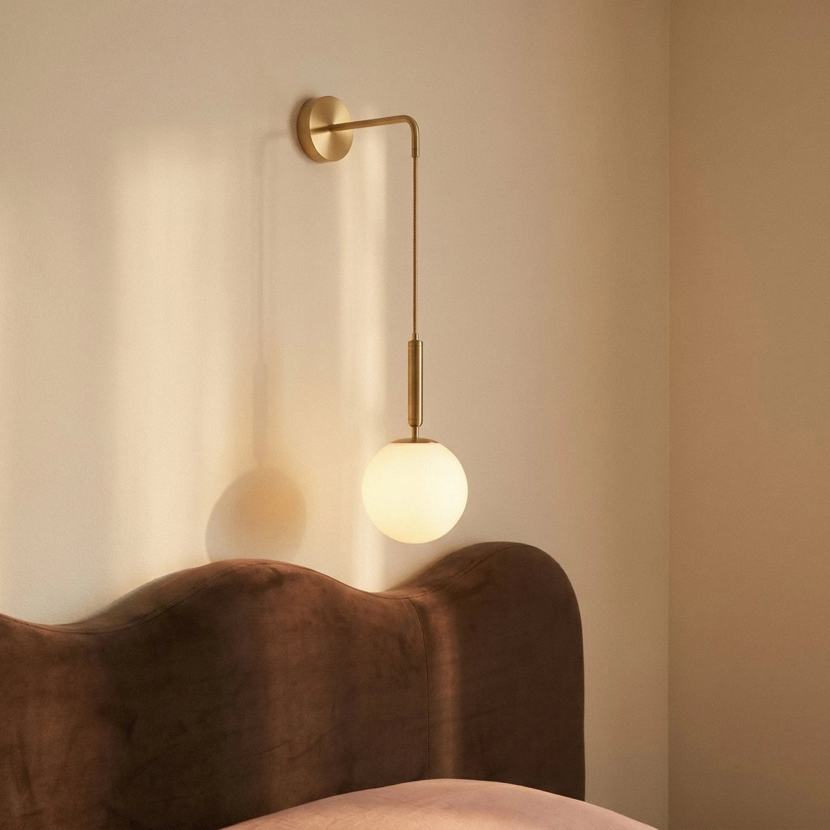 Clérachic gold globe wall sconce glowing beside a brown velvet headboard, its cord dropping to an opal glass globe.