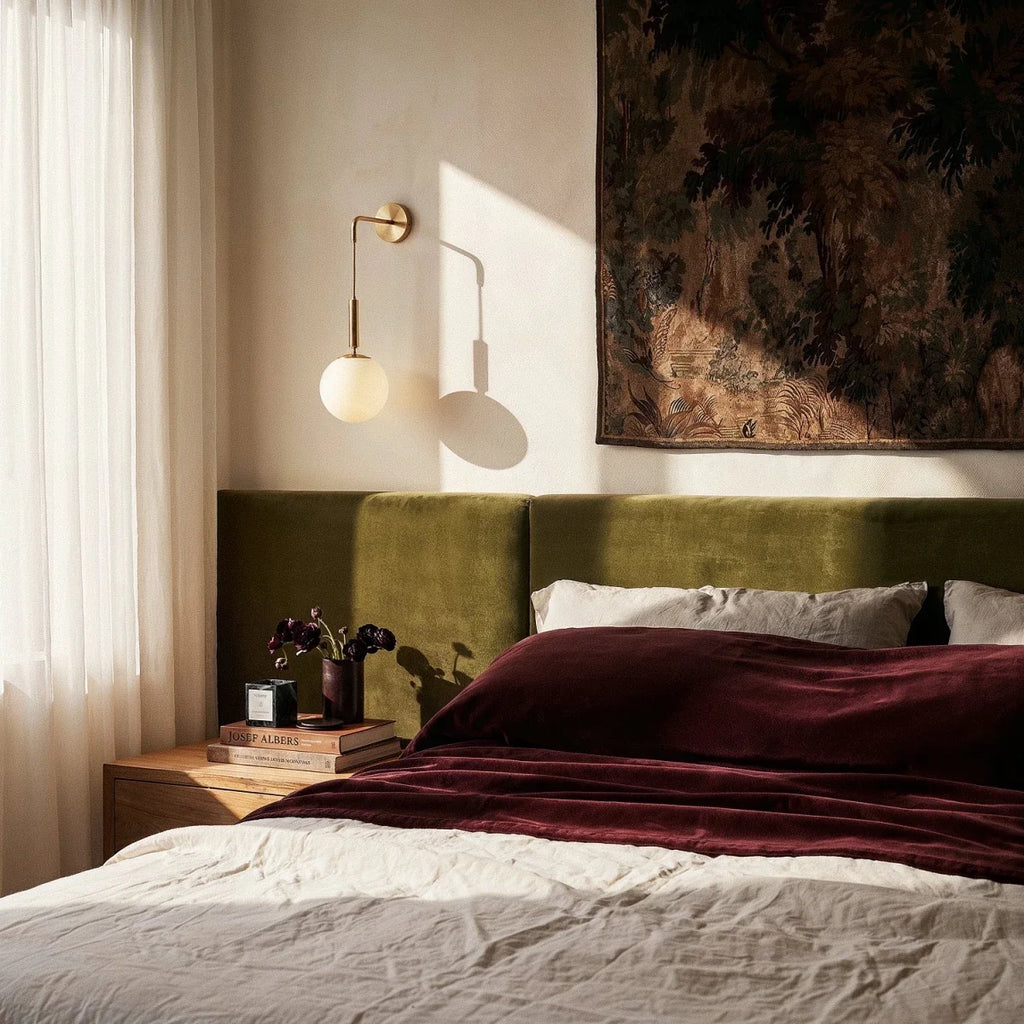Clérachic gold globe wall sconce lit above a wooden nightstand, its shadow falling across a cream bedroom wall.