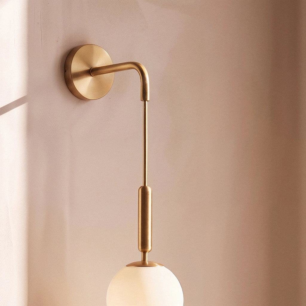 Close-up of the Clérachic gold globe sconce's brass arm, cable and opal glass globe against a blush plaster wall.