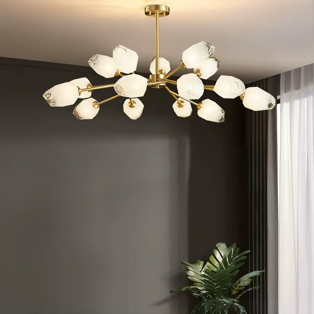 Brass arms and white faceted glass shades of this Clérachic chandelier, lit against a dark grey wall beside a potted plant.
