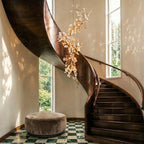 Clérachic cascading leaf chandelier hanging beside a curved wooden staircase, lit warm between tall windows.