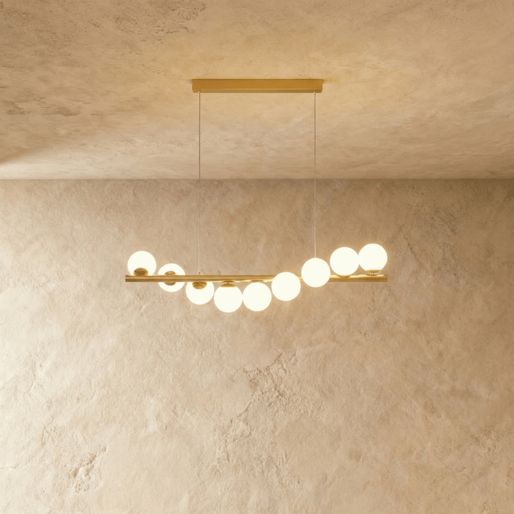 Clérachic gold linear chandelier with nine opal white glass globes on a brass bar, lit warm and suspended on two thin wires.